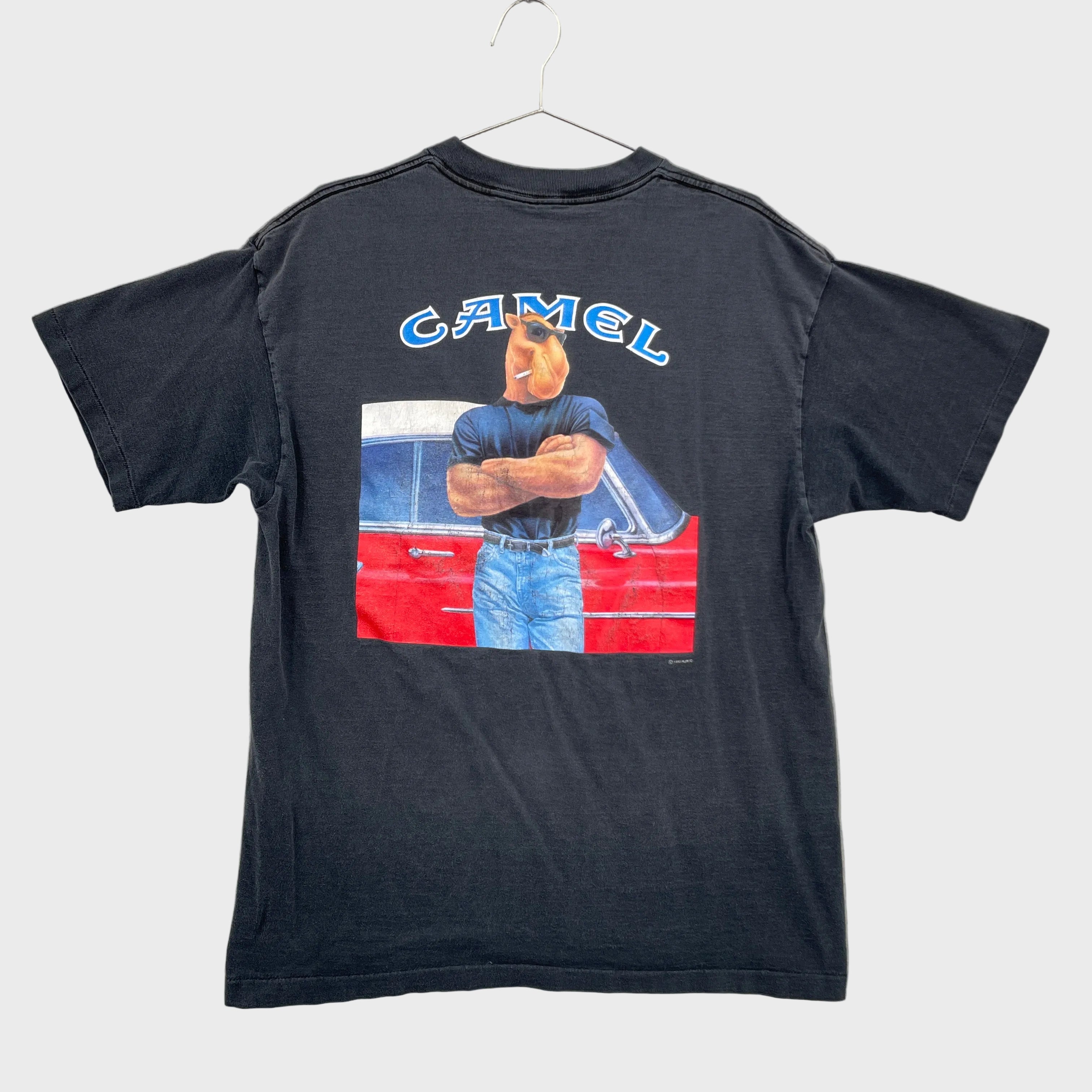 Black t-shirt with a graphic design on a gray surface. Camel Cigaretts logo with a camel character hands crossed standing in front of a vintage car