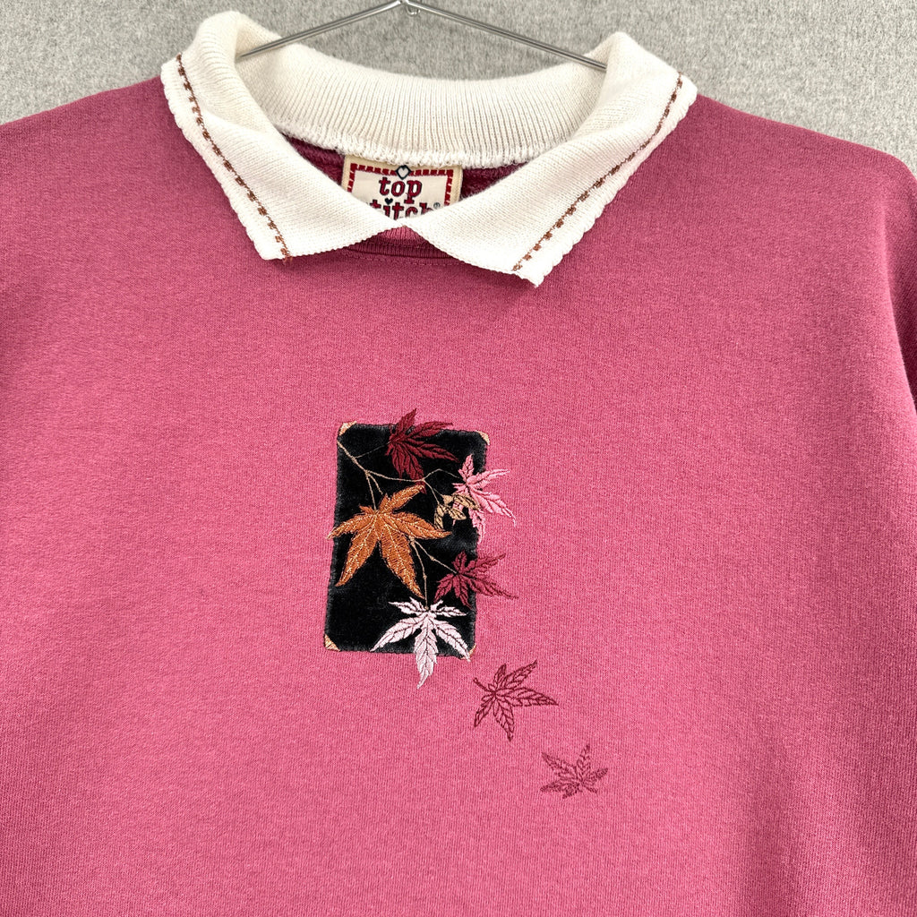 Pink sweater with embroidered design of fall leaves on a hanger against a gray background