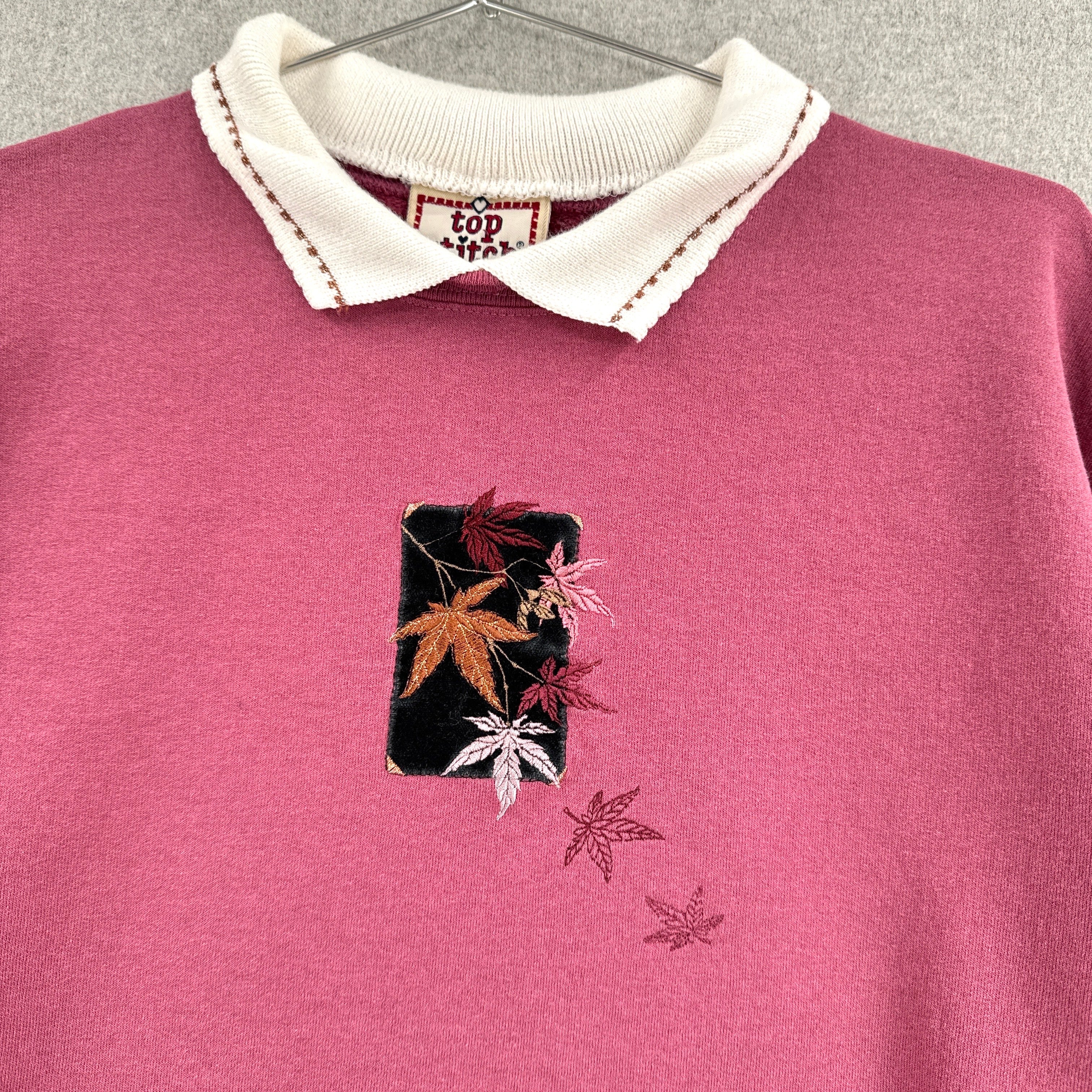 Pink sweater with embroidered design of fall leaves on a hanger against a gray background