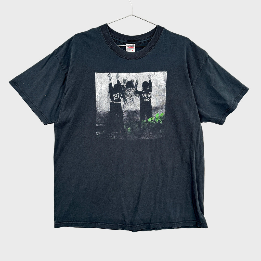 Black t-shirt with graphic design on a textured gray background/ The Clash White Riot. Graphic of 3 men with their hands up against a wall.