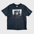 Black t-shirt with graphic design on a textured gray background/ The Clash White Riot. Graphic of 3 men with their hands up against a wall.