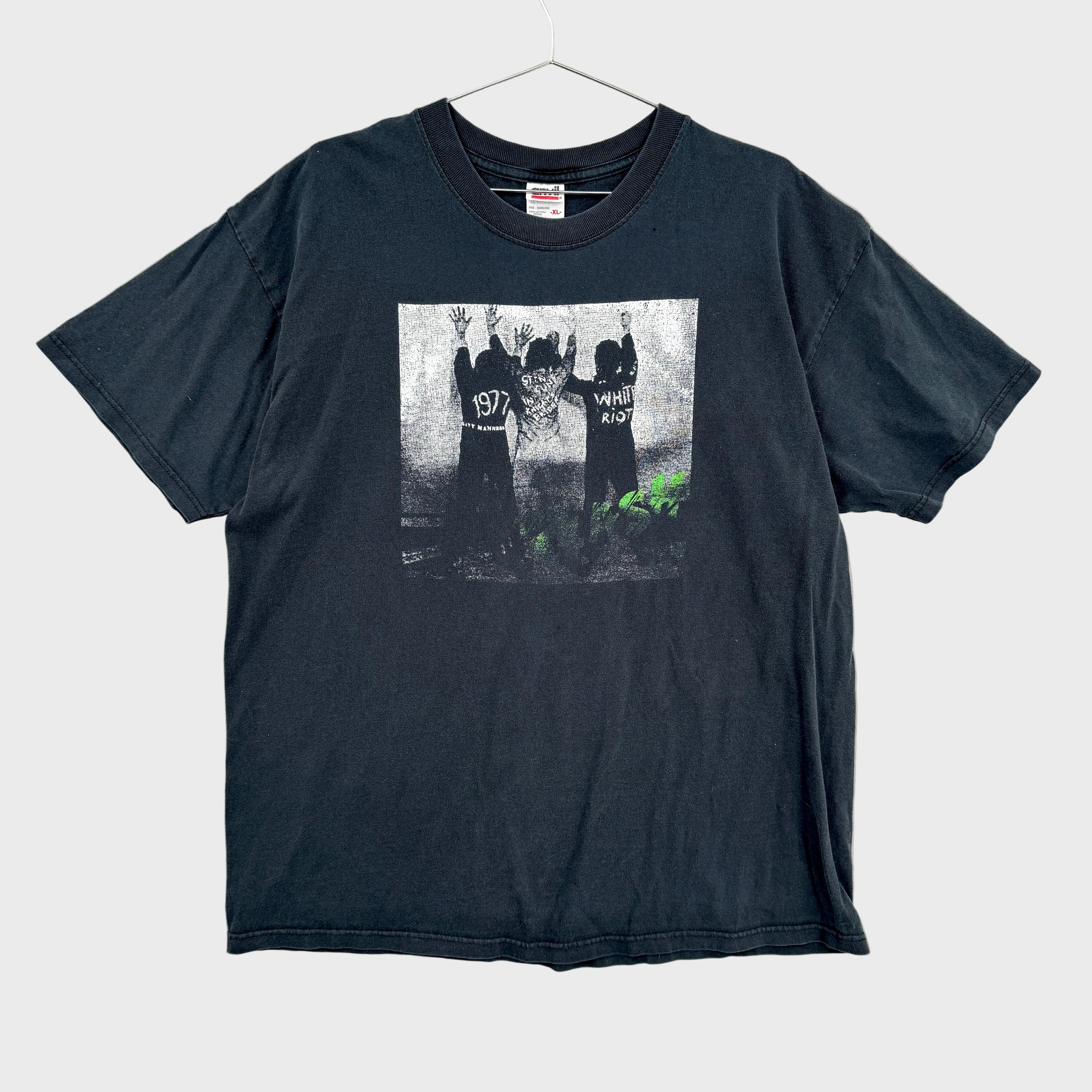 Black t-shirt with graphic design on a textured gray background/ The Clash White Riot. Graphic of 3 men with their hands up against a wall.