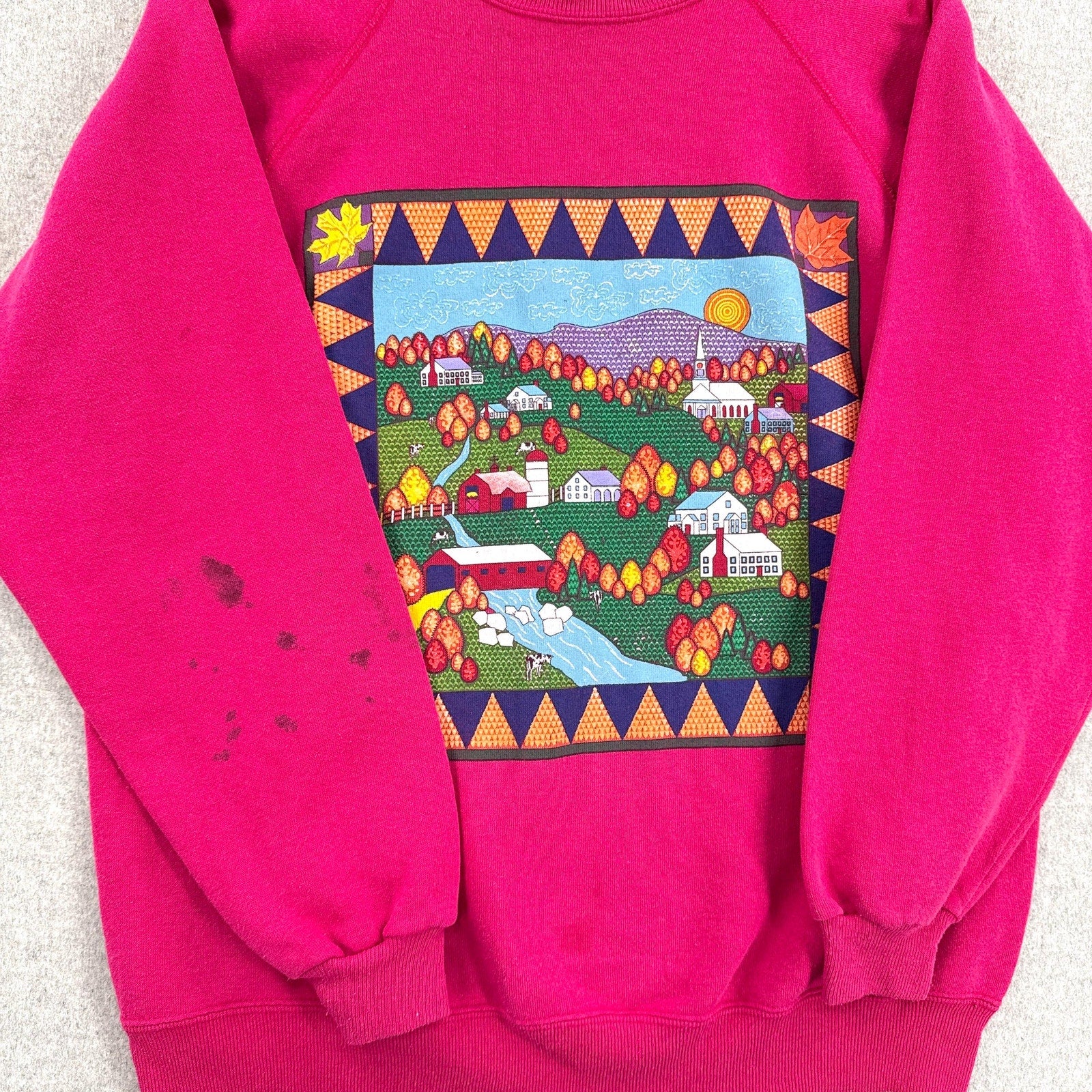 Pink sweatshirt with a colorful graphic of a village scene on a light gray background. stain spot on sleeve