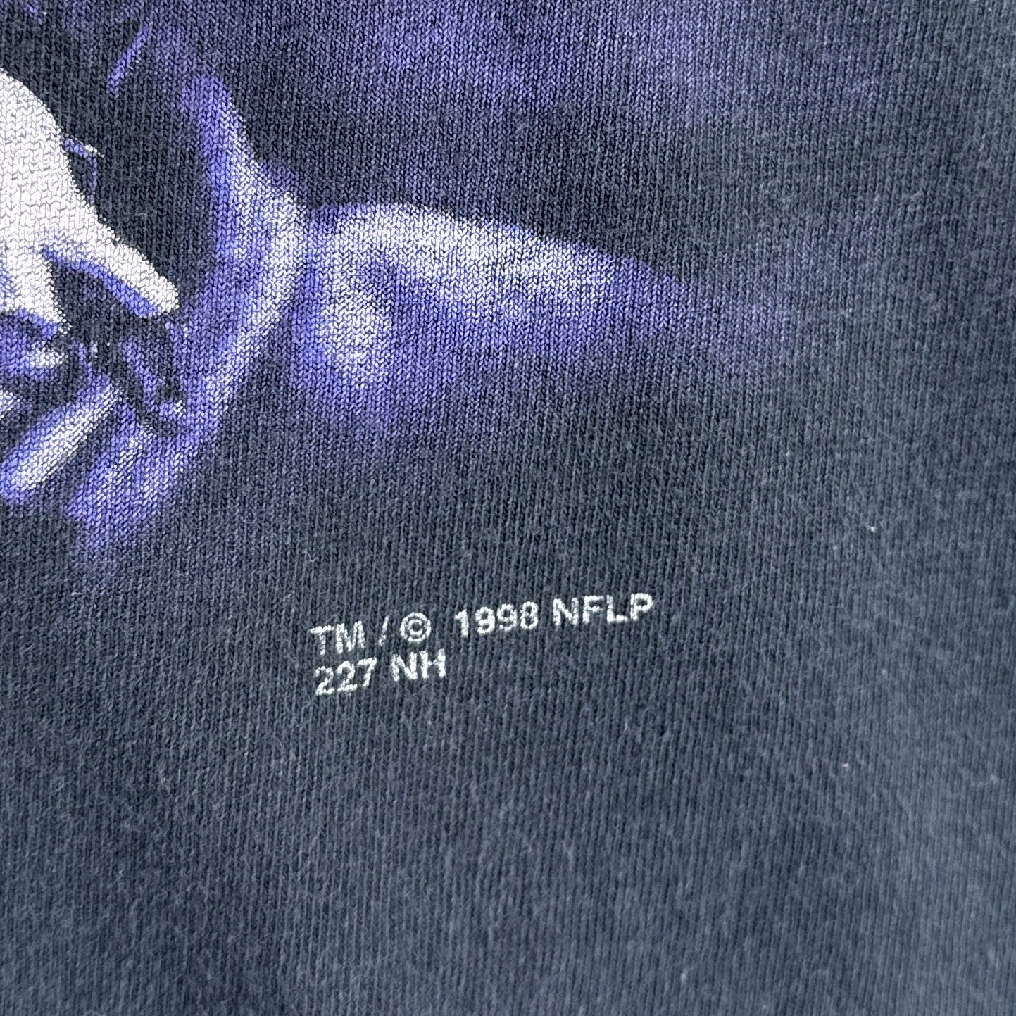 Close-up of a dark blue shirt with a logo and text, including 'TM ® 1998 NFLP' on a dark background.