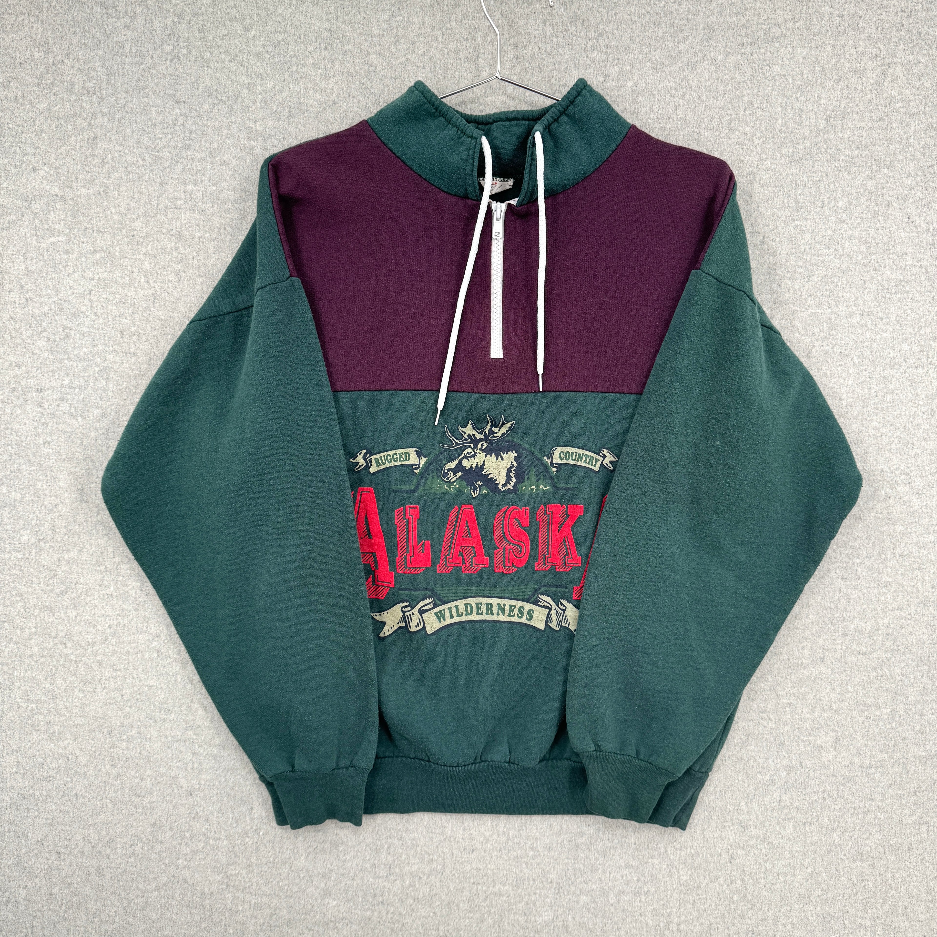 Green and purple sweatshirt with 'Alaska' graphic on a gray background. Sleeves folded over not displaying any defects.