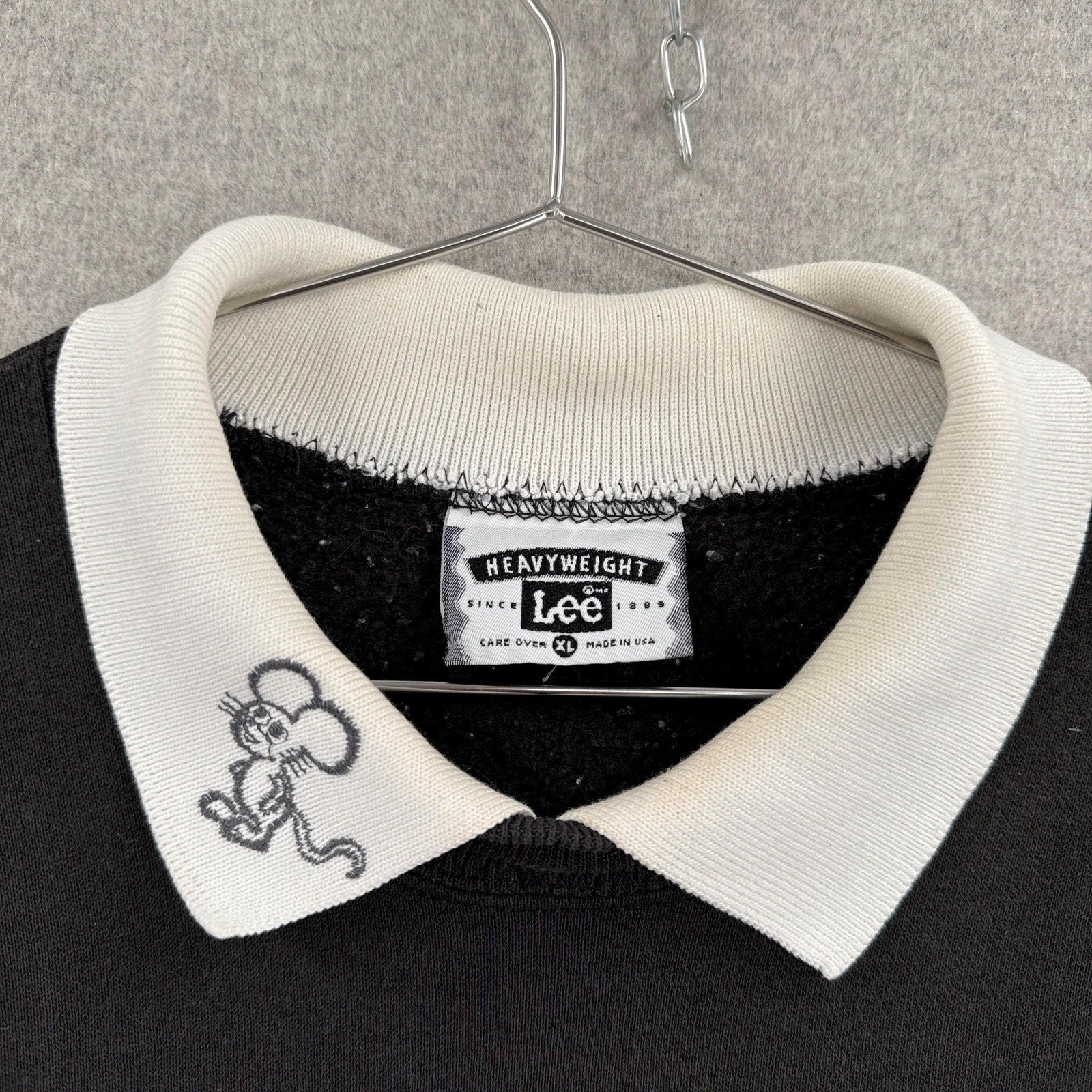 Black sweater with white collar and embroidered design on a hanger against a gray background