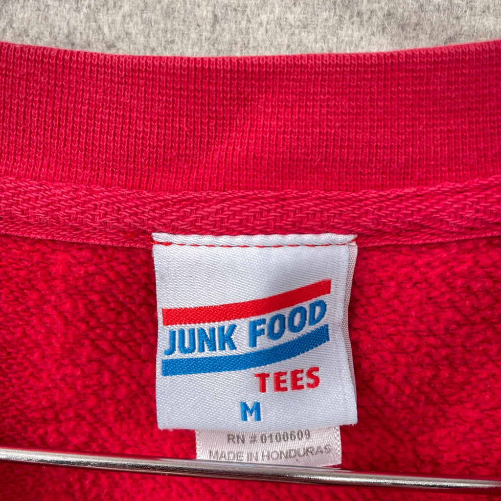 Close-up of a red garment with a 'Junk Food Tees' label on a textured gray background. Tag size Medium