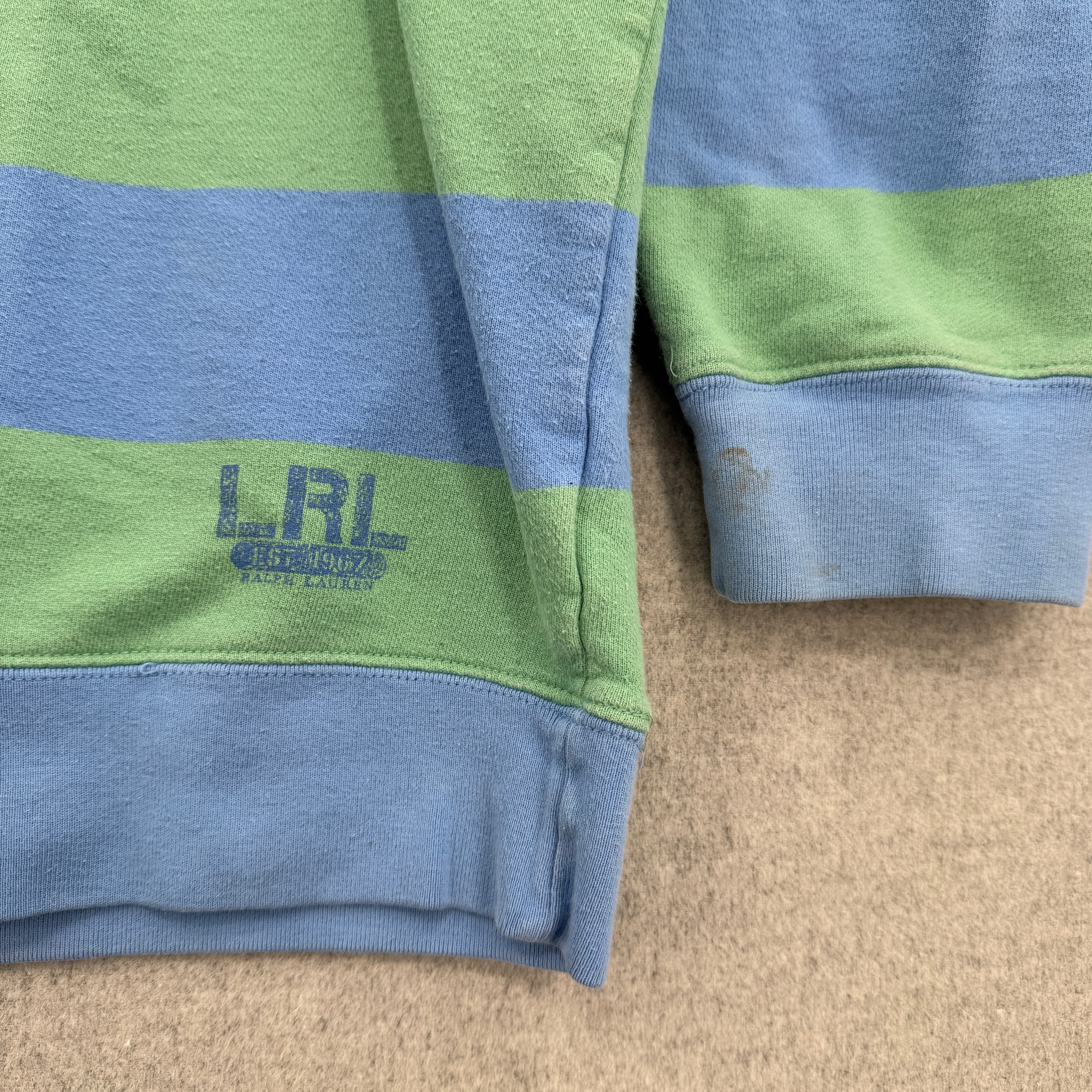 Close-up of a green and blue striped fabric with 'LRL' logo on a beige surface, stain spots on the sleeve