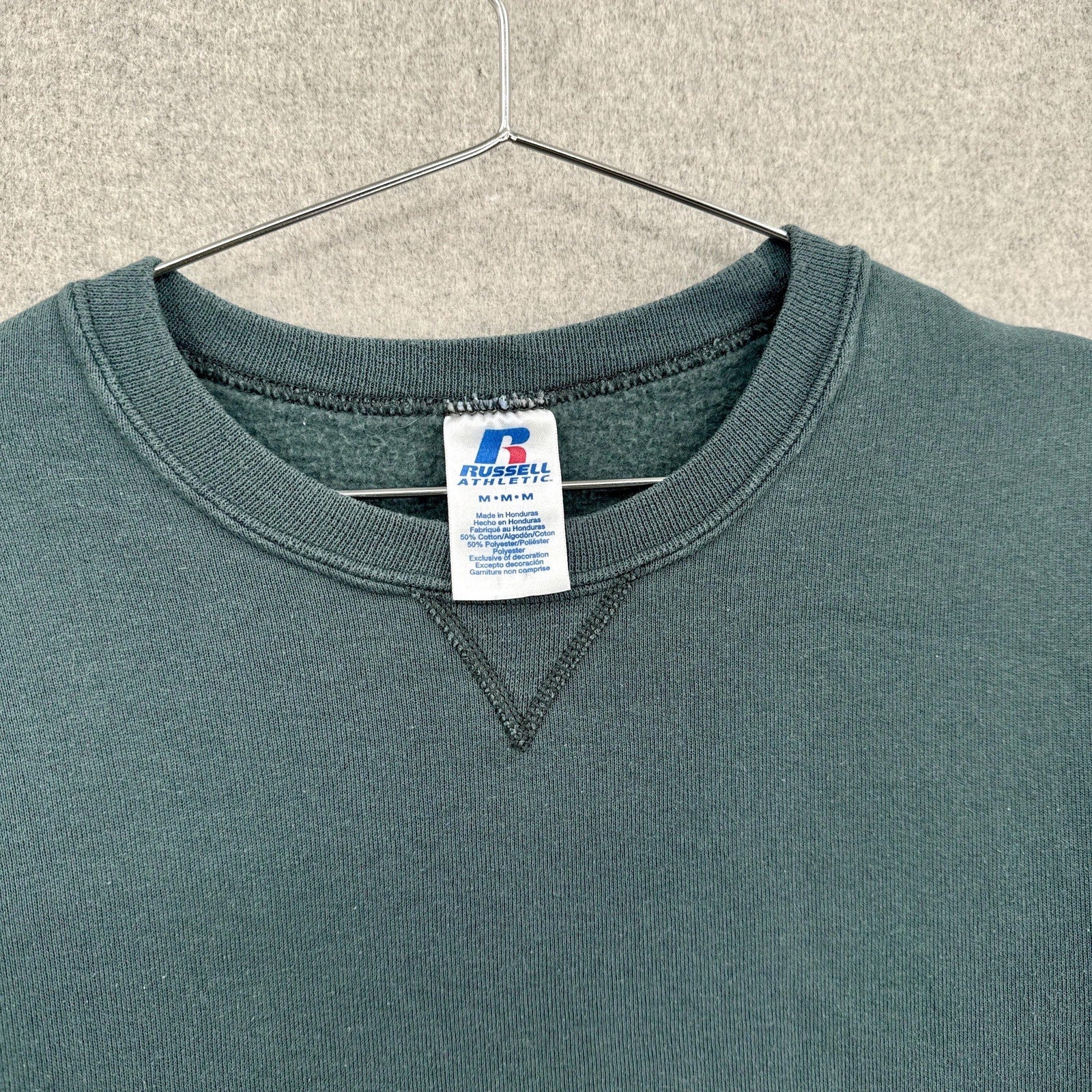 Greenish blue grey sweatshirt with a brand label on a hanger against a neutral background