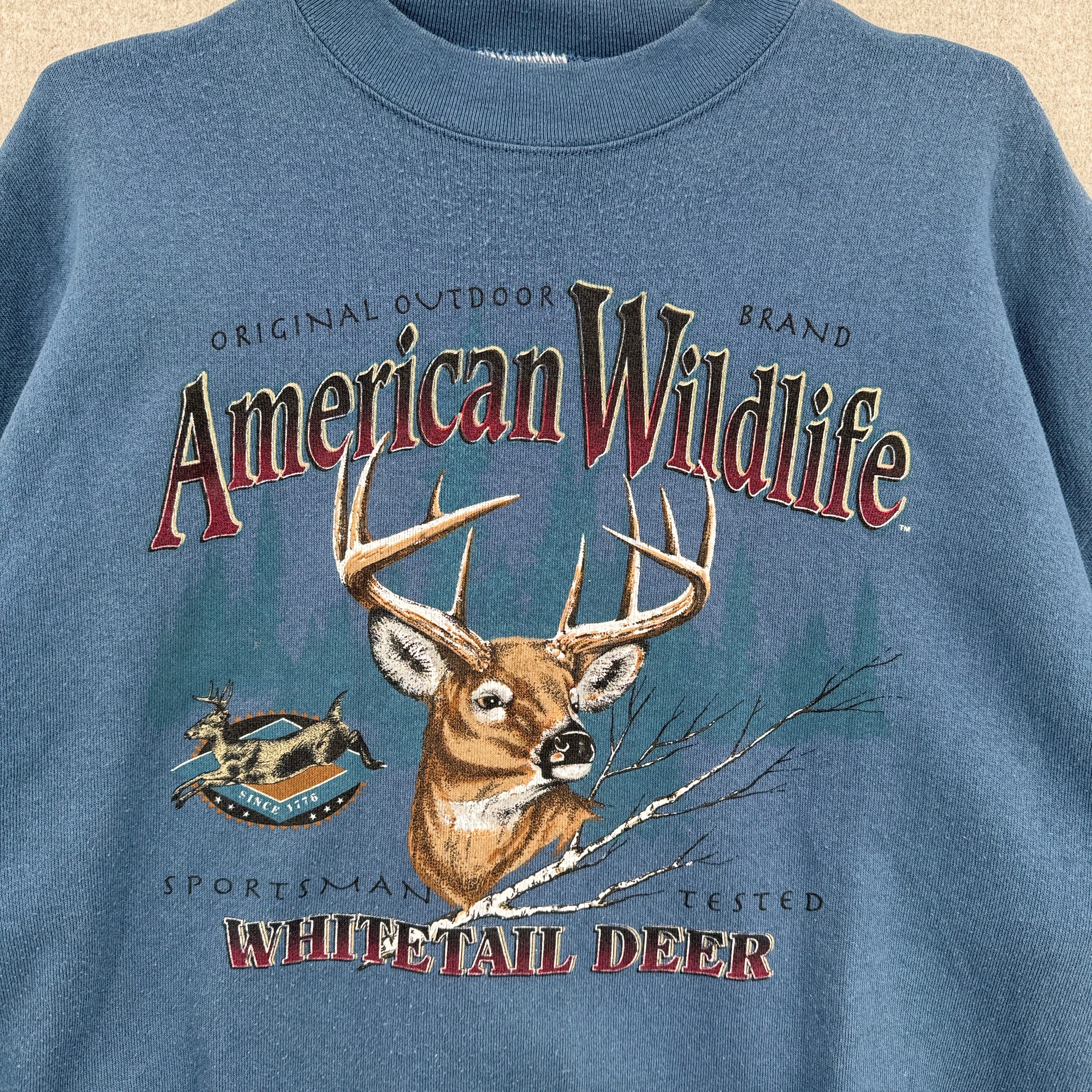Blue sweatshirt with 'American Wildlife' and 'Whitetail Deer' graphic on a beige background