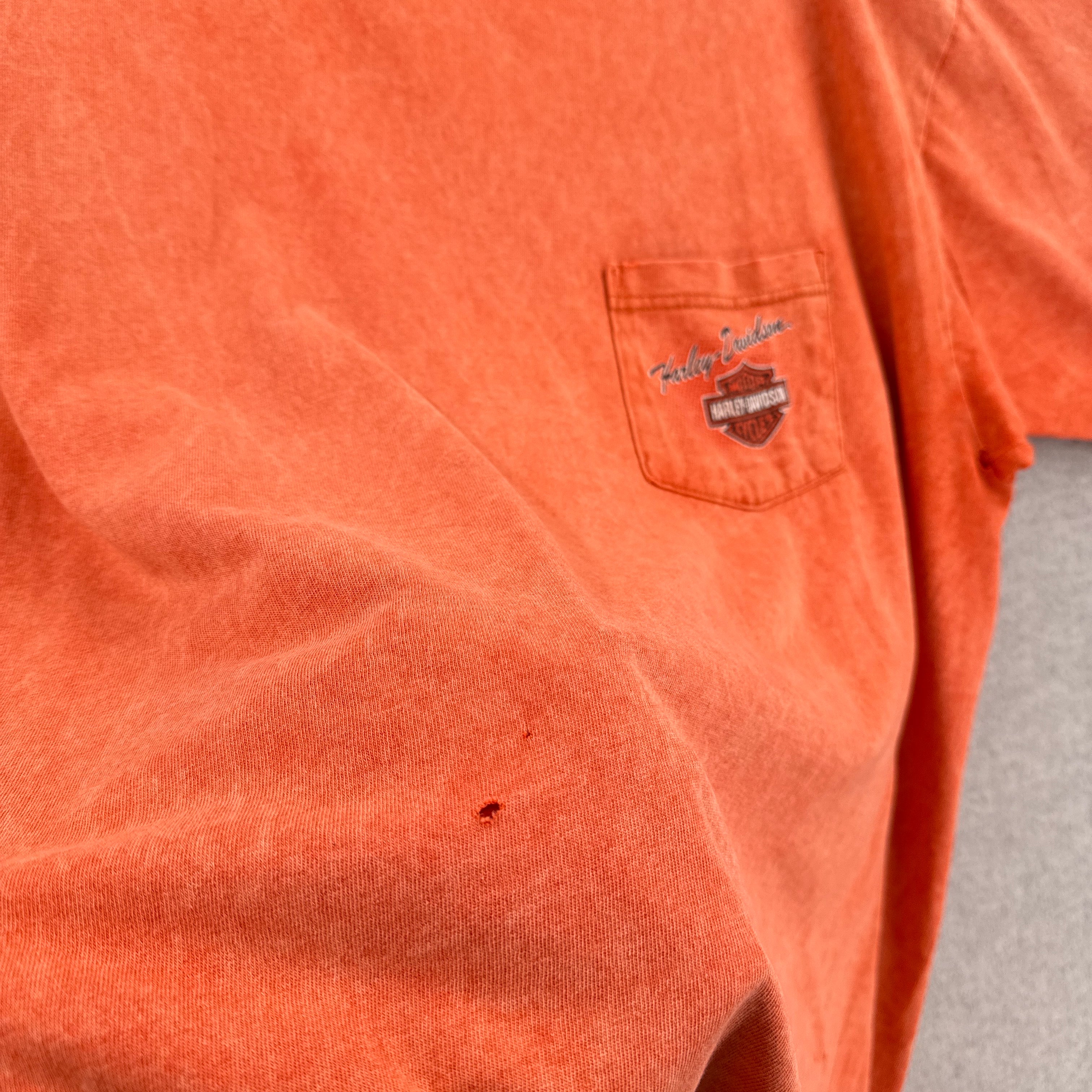 Close-up of an orange shirt with a logo on the chest pocket against a gray background. small hole on the front