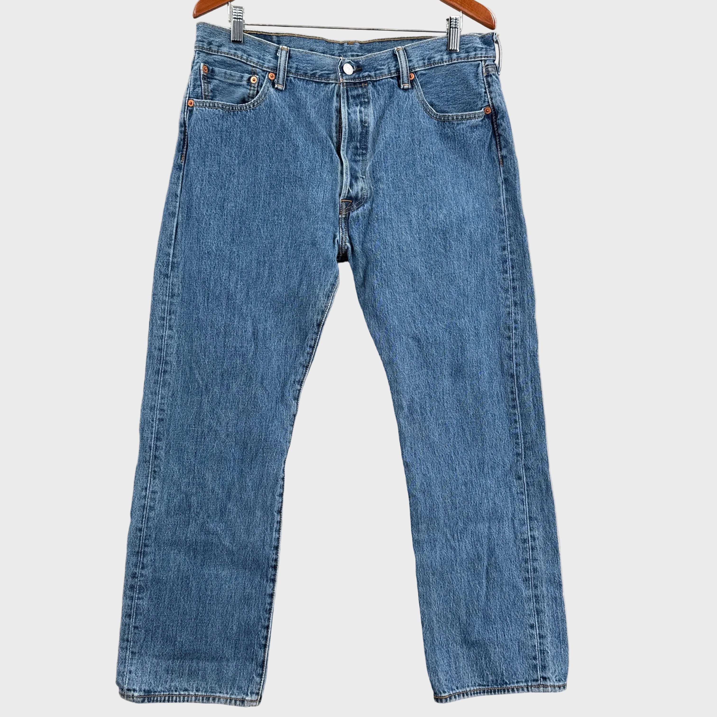 Pair of blue jeans hanging on a wooden hanger against a beige wall.