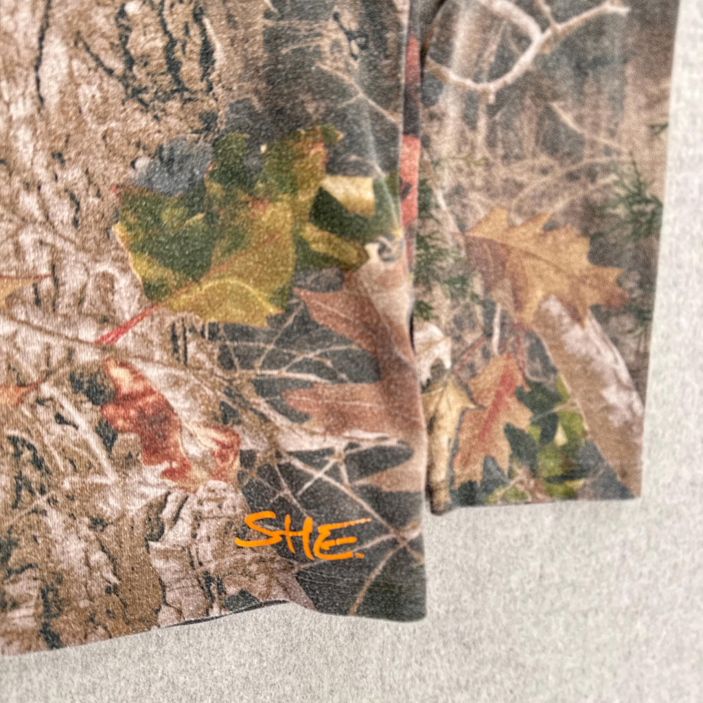 Close-up of camouflage-patterned fabric with 'SHE' logo on a neutral background