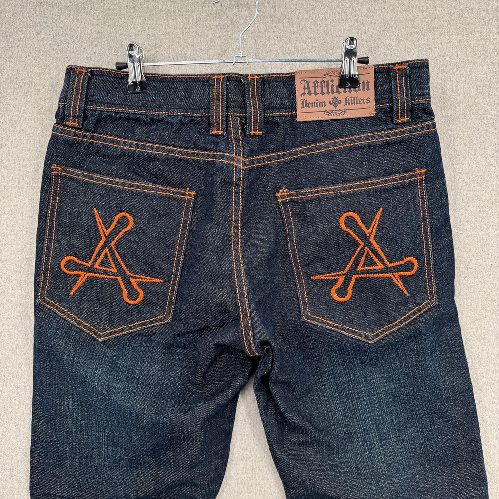 Pair of blue jeans with orange embroidery on the pockets, hanging on a beige wall.