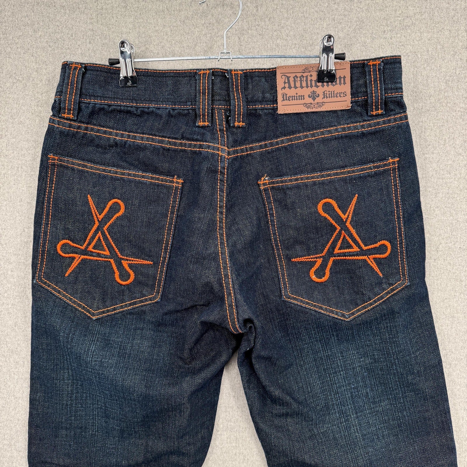 Pair of blue jeans with orange embroidery on the pockets, hanging on a beige wall.